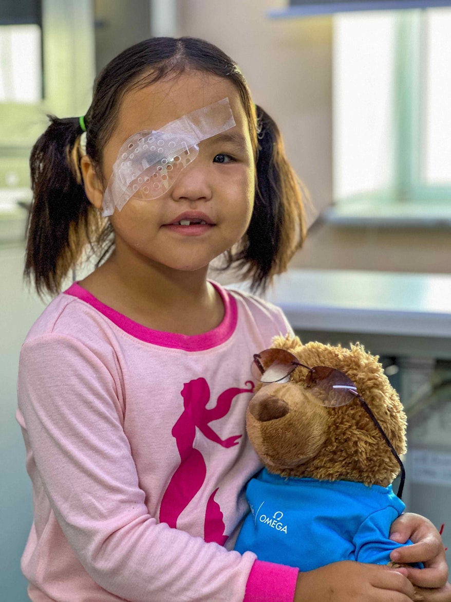 A young girl with a patch over her eye holds a teddy bear after surgery.